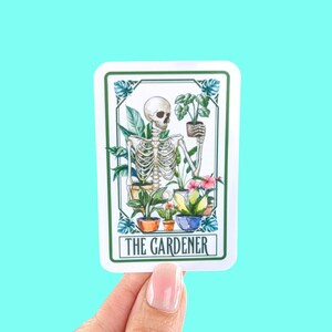 Puede incluir: Una pegatina blanca con un borde verde muestra un esqueleto sosteniendo una planta en maceta. El esqueleto est&aacute; rodeado de otras plantas en maceta. El texto "THE GARDENER" est&aacute; impreso en verde en la parte inferior de la pegatina.