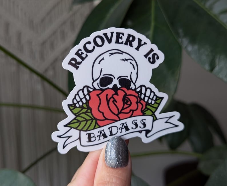 May include: White sticker with a black and white skull holding a red rose with green leaves. The sticker has the text "Recovery is Badass" in black.