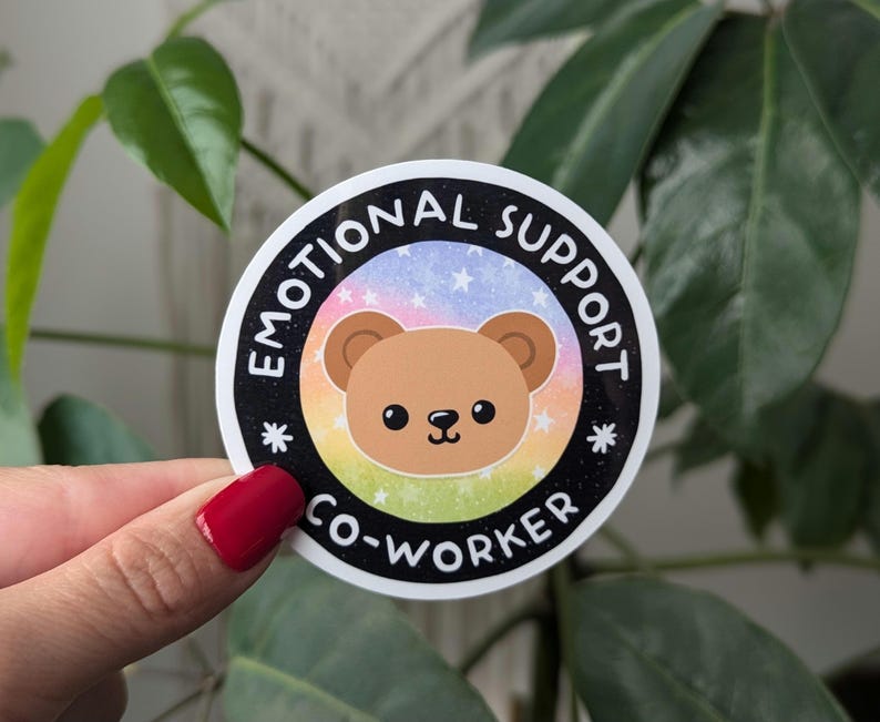 K&ouml;nnte beinhalten: Ein runder Aufkleber mit schwarzem Hintergrund und wei&szlig;em Text, der "Emotional Support Co-Worker" lautet. Der Aufkleber zeigt einen Comic-B&auml;ren mit braunem Fell und einem Regenbogenhintergrund.