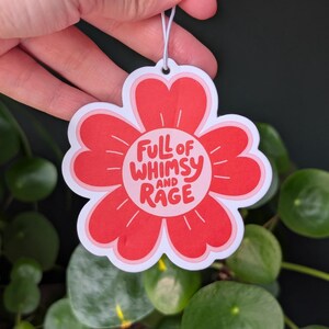 Może przedstawiać: Czerwony odświeżacz powietrza w kształcie kwiatu z białym napisem "FULL OF WHIMSY AND RAGE". Odświeżacz powietrza jest trzymany w dłoni na tle zielonych roślin.