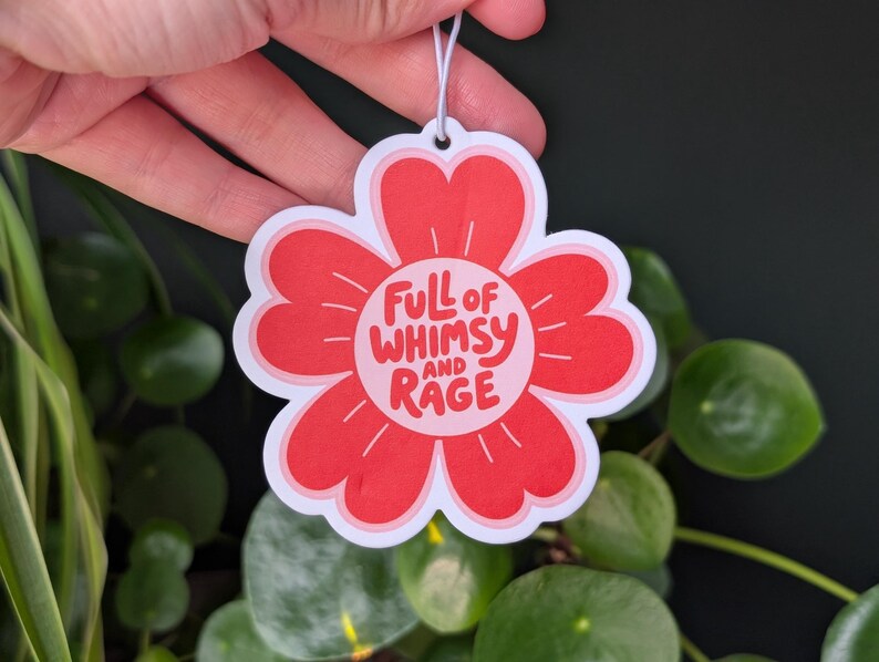 Może przedstawiać: Czerwony odświeżacz powietrza w kształcie kwiatu z białym napisem "FULL OF WHIMSY AND RAGE". Odświeżacz powietrza jest trzymany w dłoni na tle zielonych roślin.