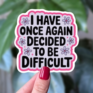 Puede incluir: Una pegatina con borde rosa y el texto "I HAVE ONCE AGAIN DECIDED TO BE DIFFICULT" en negro. La pegatina tiene un fondo blanco con detalles de flores moradas.
