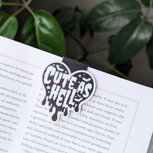 May include: A black and white heart-shaped bookmark with the text "Cute as Hell" in dripping white letters. The bookmark has a black background and white bats and stars.