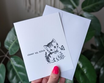 Könnte beinhalten: Eine weiße Grußkarte mit einer Schwarz-Weiß-Illustration eines Kätzchens und eines Schmetterlings, mit dem Text "cheer up, bitch". Die Karte wird von einem passenden weißen Umschlag begleitet.