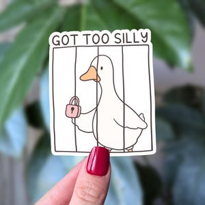 May include: A white sticker featuring a cartoon duck with an orange beak inside a black-lined cage, holding a small pink padlock. The text GOT TOO SILLY is printed in black capital letters at the top of the rectangular sticker design.