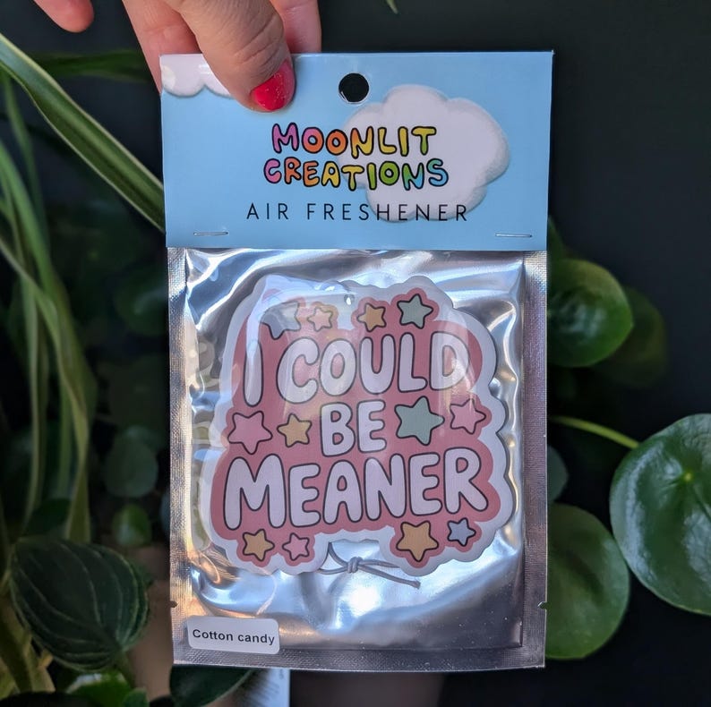 May include: An air freshener in a clear plastic package. The air freshener has a pink and white design with the words "I COULD BE MEANER" in a playful font, surrounded by stars. The packaging reads "MOONLIT CREATIONS AIR FRESHENER" and "Cotton candy".