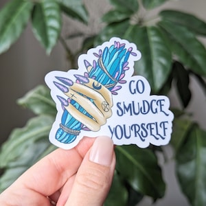 May include: A sticker with a hand holding a blue and purple smudge stick with the text "Go Smudge Yourself".