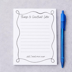 May include: A white notepad with the text "Things to Overthink Later" and "...shit I need more room" in black script. The notepad has a decorative border and lined pages. A blue pen rests beside the notepad.