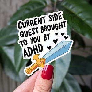 Op de afbeelding: Een sticker met een blauw zwaard en de tekst "Current side quest brought to you by ADHD" met hartjes rond de tekst.