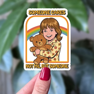 May include: A die-cut sticker featuring a cartoon illustration of a smiling child with brown hair wearing a yellow shirt with a mushroom pattern, holding a brown teddy bear. A rainbow arches behind them. The text reads SOMEONE CARES NOT ME, BUT SOMEONE.