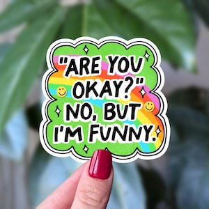 May include: A colourful sticker with the text "ARE YOU OKAY?" "NO, BUT I'M FUNNY." The sticker has a green cloud-like border with rainbow accents and smiley faces. The sticker is held by a hand with red nail varnish.