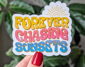 May include: A white sticker with a colorful graphic design. The sticker features the text "FOREVER CHASING SUNSETS" in a retro font with yellow, pink, and blue colors. A stylized sun with yellow rays is above the text.