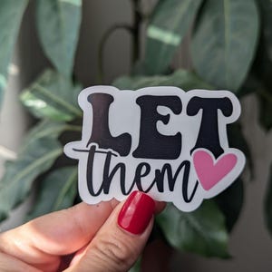 Op de afbeelding: Een witte sticker met de woorden "LET them" in een zwarte, vette letter, met een roze hart. De sticker wordt voor een achtergrond van groene bladeren gehouden.