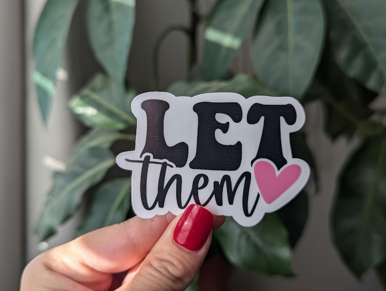 Op de afbeelding: Een witte sticker met de woorden "LET them" in een zwarte, vette letter, met een roze hart. De sticker wordt voor een achtergrond van groene bladeren gehouden.
