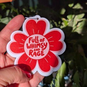 Może przedstawiać: Czerwono-biała naklejka w kształcie kwiatu z napisem "Full of Whimsy and Rage" w zabawnej czcionce. Naklejka ma mały otw&oacute;r u g&oacute;ry do zawieszenia. Tło to mieszanka zielonych liści.