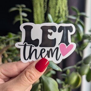 Op de afbeelding: Een witte sticker met de woorden "LET them" in zwarte, gestileerde letters. Een roze hart staat naast het woord "them". De sticker wordt vastgehouden in een hand met rode nagellak, met een achtergrond van groene planten.