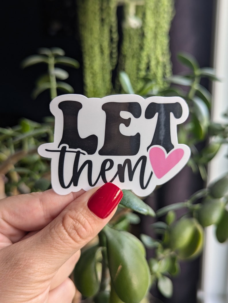 Op de afbeelding: Een witte sticker met de woorden "LET them" in zwarte, gestileerde letters. Een roze hart staat naast het woord "them". De sticker wordt vastgehouden in een hand met rode nagellak, met een achtergrond van groene planten.