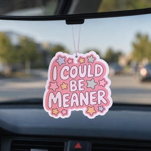 May include: A pink car air freshener with the text "I COULD BE MEANER" in a playful font, surrounded by star and cloud shapes. The air freshener hangs from a car's rearview mirror, with a blurred street scene in the background.