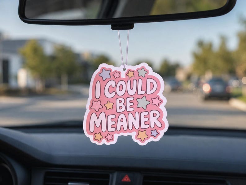 May include: A pink car air freshener with the text "I COULD BE MEANER" in a playful font, surrounded by star and cloud shapes. The air freshener hangs from a car's rearview mirror, with a blurred street scene in the background.