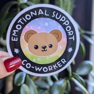 K&ouml;nnte beinhalten: Ein runder Aufkleber mit schwarzem Rand und wei&szlig;em Hintergrund. Der Aufkleber zeigt einen braunen Comic-B&auml;ren mit einem Regenbogen- und Sternenhintergrund. Der Text "Emotional Support Co-Worker" ist in einem schwarzen Kreis um den B&auml;ren geschrieben.