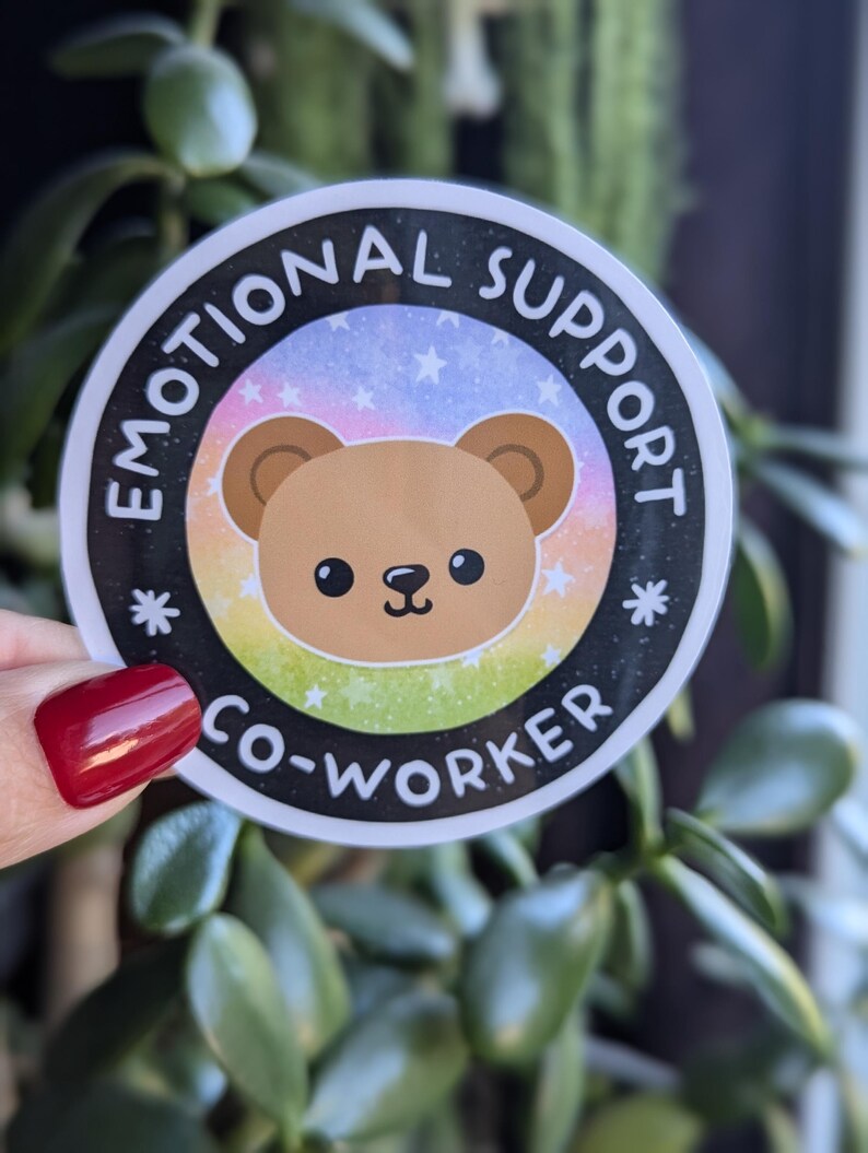 K&ouml;nnte beinhalten: Ein runder Aufkleber mit schwarzem Rand und wei&szlig;em Hintergrund. Der Aufkleber zeigt einen braunen Comic-B&auml;ren mit einem Regenbogen- und Sternenhintergrund. Der Text "Emotional Support Co-Worker" ist in einem schwarzen Kreis um den B&auml;ren geschrieben.