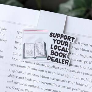 May include: A black and white sticker with the text "Support Your Local Book Dealer" in bold white letters. The sticker features a simple line drawing of an open book.