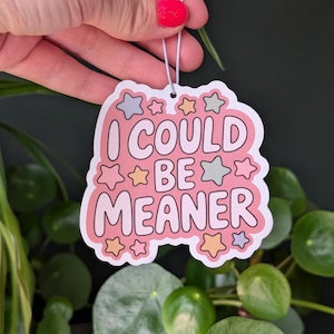 May include: A pink sticker with the text "I COULD BE MEANER" in a playful font, surrounded by star-shaped accents in pastel colors. The sticker is held by a hand with red nail polish, with green plants in the background.