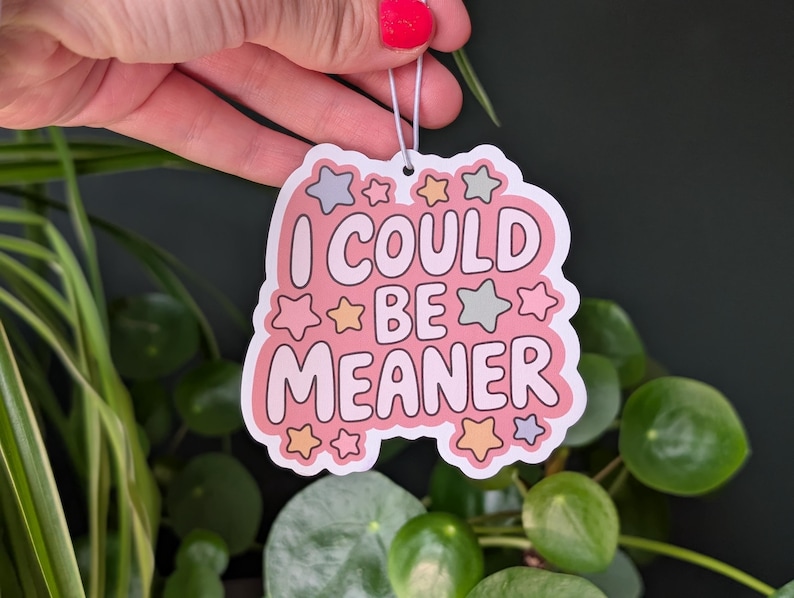 May include: A pink sticker with the text "I COULD BE MEANER" in a playful font, surrounded by star-shaped accents in pastel colors. The sticker is held by a hand with red nail polish, with green plants in the background.
