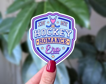 Może przedstawiać: Naklejka z jasnoniebieskim wzorem tarczy, z napisem "IN MY HOCKEY ROMANCE Era". Projekt zawiera kije hokejowe, serce i gwiazdy w kolorze różowym, niebieskim i żółtym. Naklejka jest trzymana przez dłoń z czerwonym lakierem do paznokci.