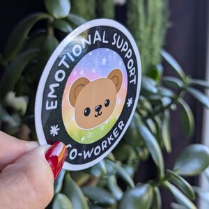 K&ouml;nnte beinhalten: Ein runder Aufkleber mit schwarzem Hintergrund und wei&szlig;em Text, der "Emotional Support Co-Worker" lautet. Der Aufkleber zeigt einen braunen Cartoon-B&auml;ren mit einem Regenbogenhintergrund und wei&szlig;en Sternen.