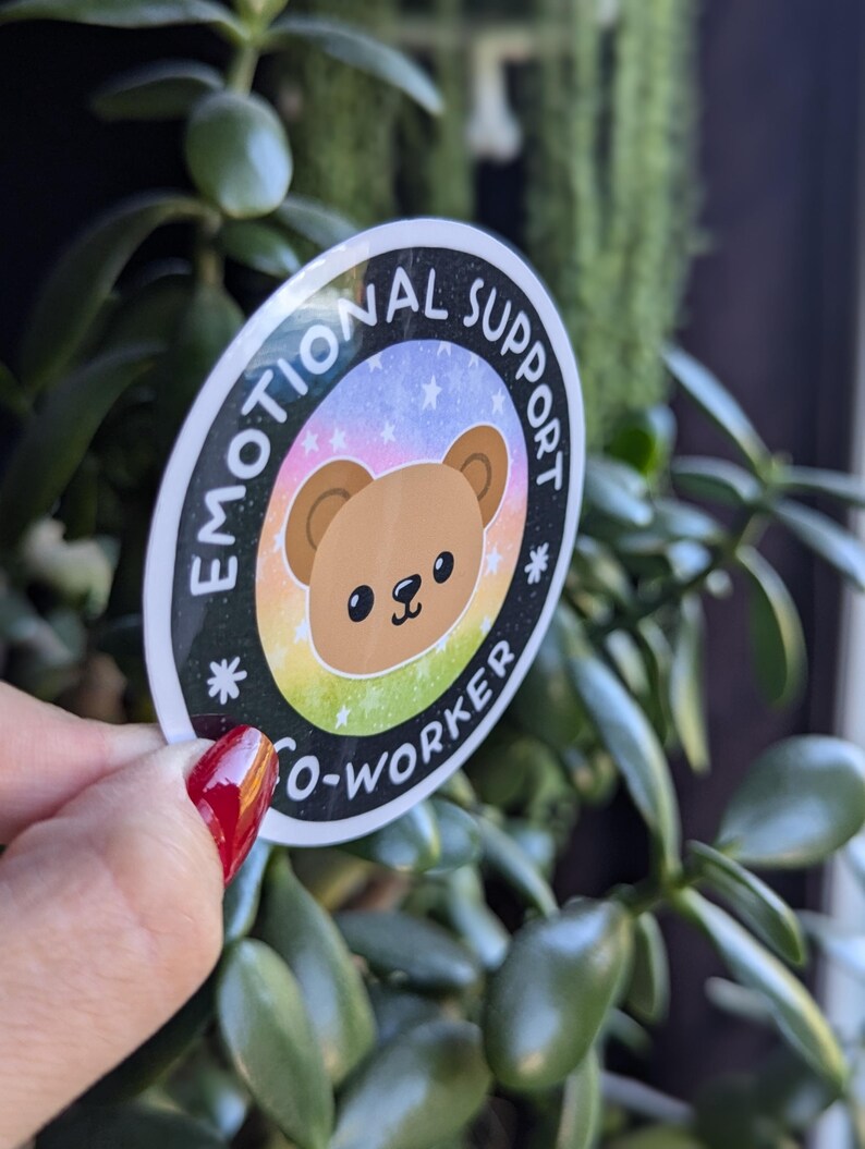 K&ouml;nnte beinhalten: Ein runder Aufkleber mit schwarzem Hintergrund und wei&szlig;em Text, der "Emotional Support Co-Worker" lautet. Der Aufkleber zeigt einen braunen Cartoon-B&auml;ren mit einem Regenbogenhintergrund und wei&szlig;en Sternen.