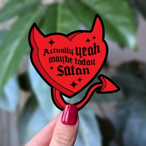 May include: A red heart-shaped sticker with devil horns and a tail. The sticker features the text "Actually yeah maybe today Satan" in black gothic font. Four small star shapes are also included in the design.