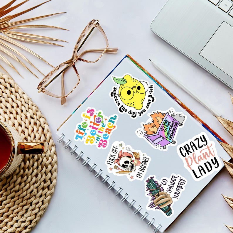 May include: A notebook with several colorful stickers featuring humorous phrases and illustrations. Stickers include a lemon with glasses, a skull baking, and the text "Crazy Plant Lady." Also visible are eyeglasses, a cup of tea, and a laptop.