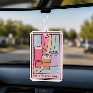 May include: A rectangular car air freshener featuring a tarot card design with a pastel rainbow background. The illustration shows a pink tumbler, a brown iced coffee with whipped cream, and a purple soda can. The text reads III and THREE OF CUPS.