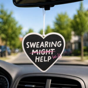 Peut inclure: Un désodorisant de voiture en forme de cœur avec un fond noir et un texte blanc indiquant SWEARING MIGHT HELP. Le mot MIGHT est barré d'une ligne rose, et le mot WILL est écrit en rose à côté, suspendu par une ficelle blanche.