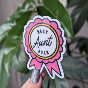 May include: A pink and yellow sticker with a white circle and the text "Best Aunt Ever" inside. The sticker is shaped like a ribbon.