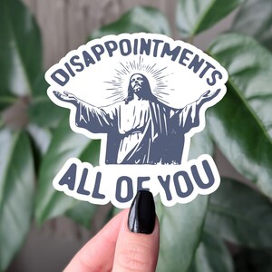 May include: A white sticker featuring a blue illustration of Jesus with open arms. The text above reads "DISAPPOINTMENTS" and below "ALL OF YOU", in a bold, sans-serif font. The sticker is held against a background of green leaves.