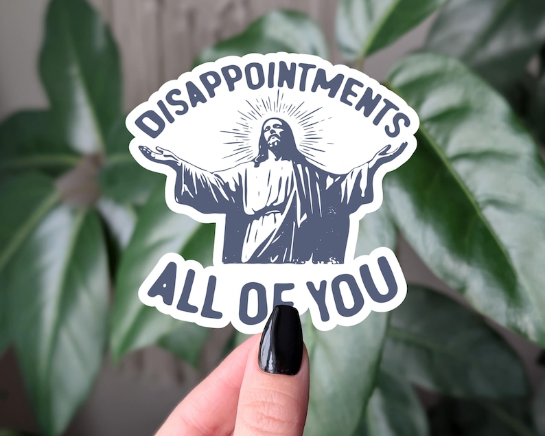 May include: A white sticker featuring a blue illustration of Jesus with open arms. The text above reads "DISAPPOINTMENTS" and below "ALL OF YOU", in a bold, sans-serif font. The sticker is held against a background of green leaves.