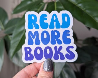 May include: A white sticker with the words "READ MORE BOOKS" in blue and purple bubble letters. The sticker is held in front of a green leafy background.