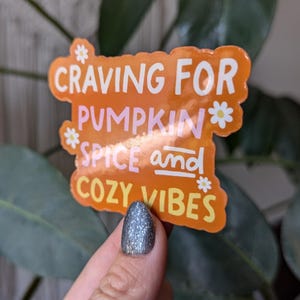 May include: Orange sticker with the text "Craving for Pumpkin Spice and Cozy Vibes" with white daisy flowers around the text.
