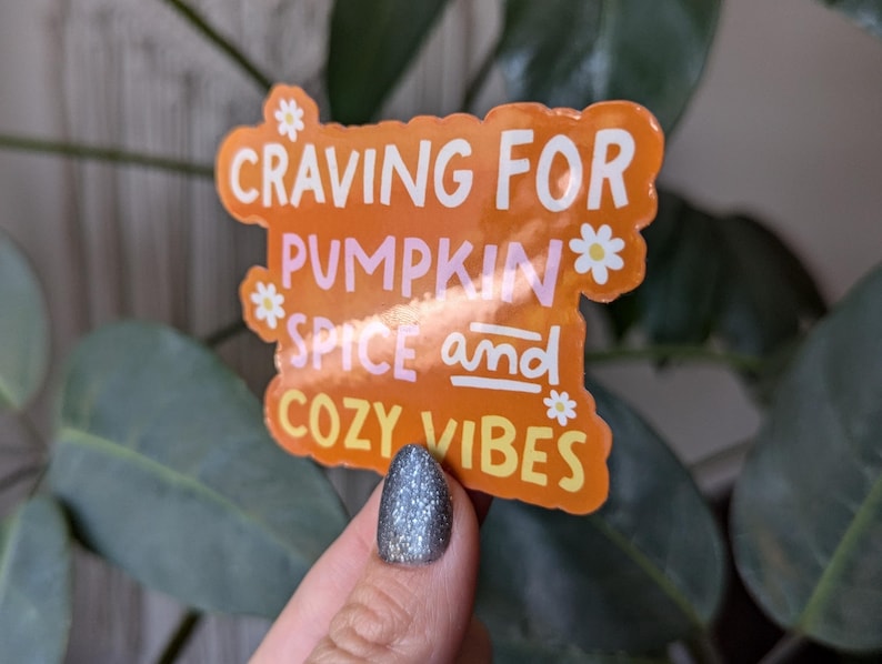 May include: Orange sticker with the text "Craving for Pumpkin Spice and Cozy Vibes" with white daisy flowers around the text.