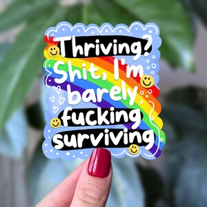 May include: A sticker with a light blue scalloped border and the text "Thriving? Shit, I'm barely fucking surviving" in black, white, and rainbow colours. Smiley faces and hearts are scattered throughout the design.