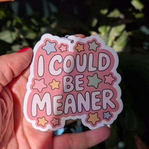 May include: A pink sticker with the text "I COULD BE MEANER" in white, outlined in pink. The sticker is decorated with star shapes in various colors, including pink, yellow, and green. A small metal ring is attached at the top.