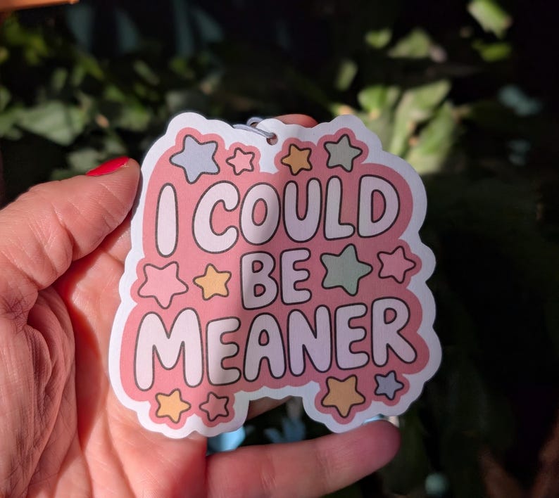 May include: A pink sticker with the text "I COULD BE MEANER" in white, outlined in pink. The sticker is decorated with star shapes in various colors, including pink, yellow, and green. A small metal ring is attached at the top.