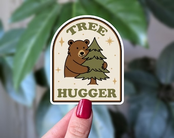 May include: A sticker with a brown bear hugging a green pine tree. The sticker has a cream-colored background with the words "TREE HUGGER" in green. Gold stars and dots surround the bear and tree.