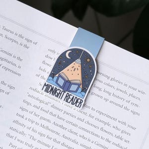 May include: A bookmark with a blue tab is inserted into a book. The bookmark features a design with a crescent moon, stars, and a book with light shining on it. The text "MIDNIGHT READER" is printed on the bookmark.
