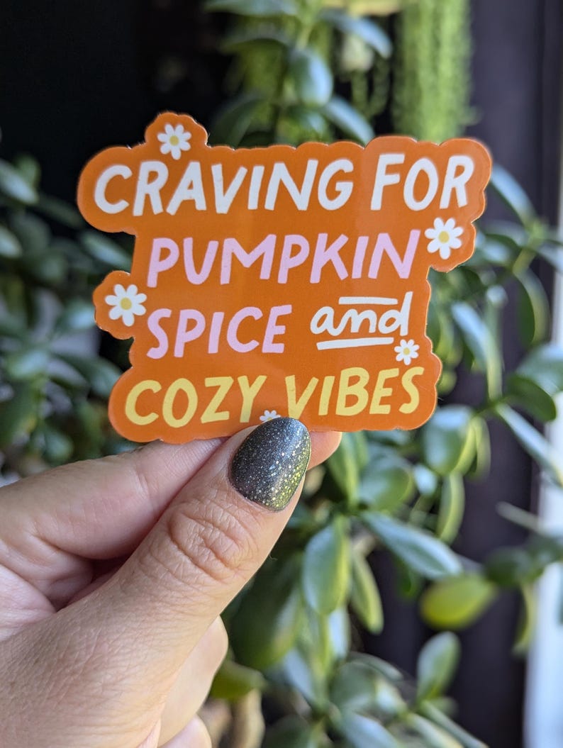 May include: Orange sticker with white text that reads "Craving for Pumpkin Spice and Cozy Vibes". The sticker has small white flower accents.