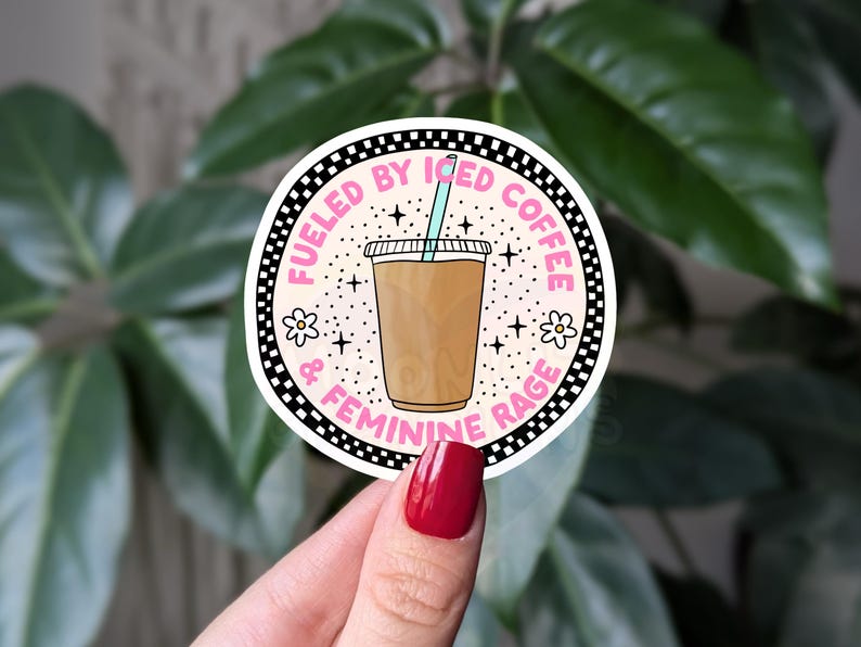 Op de afbeelding: Een ronde sticker met een roze en witte achtergrond. De sticker heeft een zwart-witte geruite rand en de tekst "Fueled by Iced Coffee & Feminine Rage". In het midden van de sticker staat een cartoonillustratie van een glas ijskoffie met een rietje.