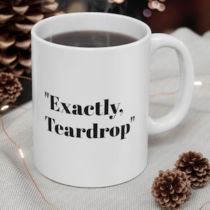 May include: White ceramic coffee mug with black text that reads "Exactly, Teardrop". The mug is filled with coffee and steam is rising from the mug. The mug is sitting on a white surface with a string of lights and pine cones.