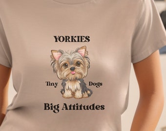 Op de afbeelding: Een beige T-shirt met een cartoon Yorkshire Terrier die een roze strik draagt. De tekst "Yorkies Tiny Dogs Big Attitudes" staat op het T-shirt gedrukt.